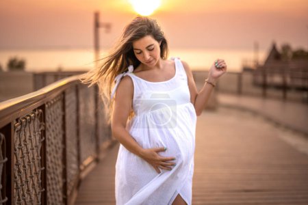 Téléchargez les photos : Portrait de femme enceinte touchant son ventre sur une robe blanche sur le trottoir de la plage au coucher du soleil - en image libre de droit