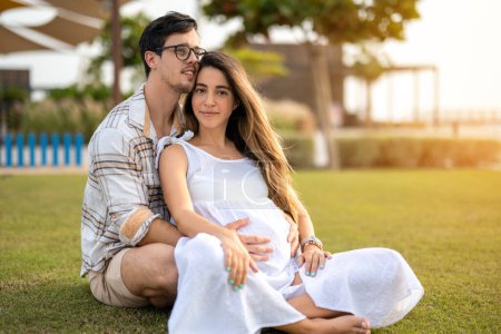 Téléchargez les photos : Belle femme enceinte et beau jeune homme assis sur l'herbe dans le parc à l'extérieur - en image libre de droit