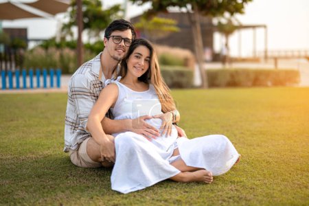 Photo for Pregnant couple woman and husband making heart on belly while sitting on grass in park - Royalty Free Image