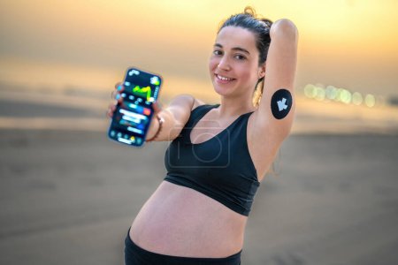 Téléchargez les photos : Belle femme enceinte utilisant Continuous Glucose Monitor pour améliorer le contrôle du diabète. Femme enceinte sportive montrant téléphone affichant test de glycémie sur smartphone App. - en image libre de droit
