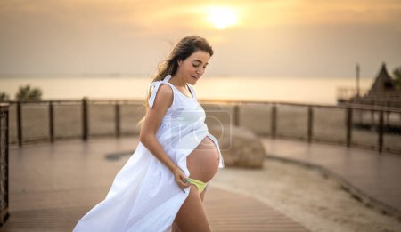 Foto de Retrato de una hermosa mujer embarazada en vestido blanco de pie en la pasarela cerca de la costa del océano al atardecer - Imagen libre de derechos