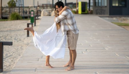 Side view of young couple hugging in romantic position while standing on wooden walkway at beach