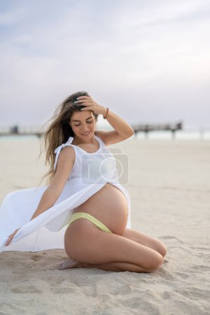 Foto de Hermosa joven embarazada sentada de rodillas y relajada con viento de brisa en la playa - Imagen libre de derechos