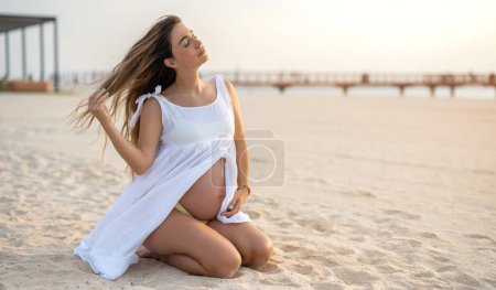 Foto de Hermosa joven embarazada sentada de rodillas disfrutando de hermosos momentos en la playa durante el atardecer - Imagen libre de derechos