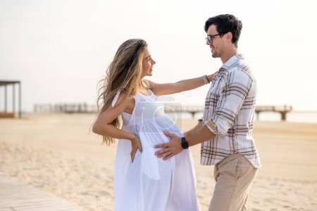 Téléchargez les photos : Beau jeune couple enceinte embrassant et regardant vers l'autre sur la plage au coucher du soleil - en image libre de droit