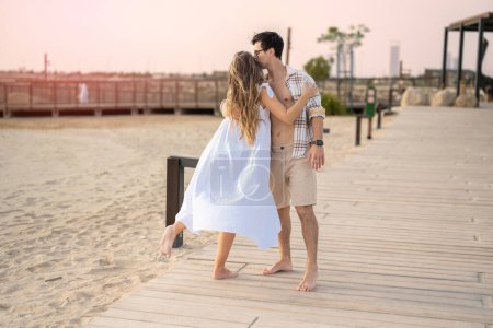 Téléchargez les photos : Beau jeune couple amoureux embrassant tout en se tenant debout sur la promenade de la plage au coucher du soleil. Beau jeune homme avec chemise ouverte embrasser sa petite amie. - en image libre de droit