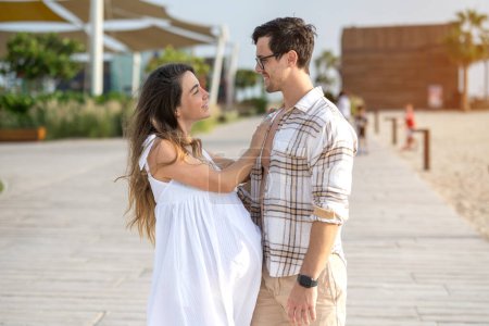 Foto de Hermosa joven pareja embarazada abrazándose y mirándose en la pasarela de la playa - Imagen libre de derechos