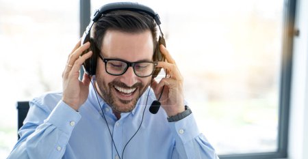 Close up portrait of cheerful handsome middle aged bearded man wearing eyeglass using headset while chatting to customer at helpdesk office.