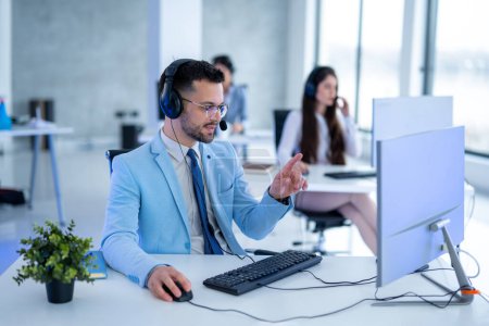 Foto de Operador masculino guapo con auriculares en traje azul que trabaja en la computadora en el centro de llamadas - Imagen libre de derechos