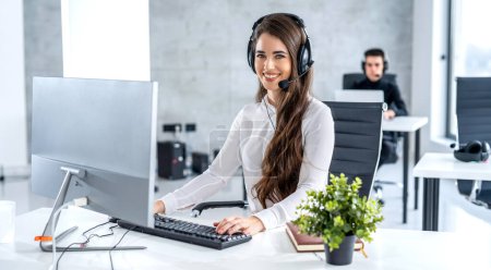 Photo for Beautiful young woman in headset working on computer and looking at camera. Telephone worker, helpdesk and customer support service concept. - Royalty Free Image