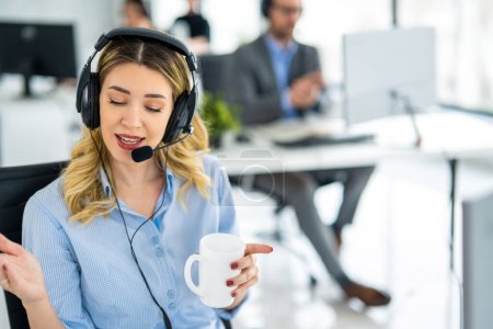 Photo for Charismatic blond hair woman wearing headset and holding coffee cup talking with customer during online phone call at helpdesk support service office. - Royalty Free Image