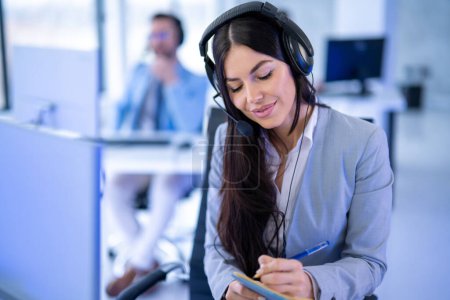 Photo for Portrait of attractive customer support service worker woman taking notes while listening customer over headset. Female operator at office. - Royalty Free Image