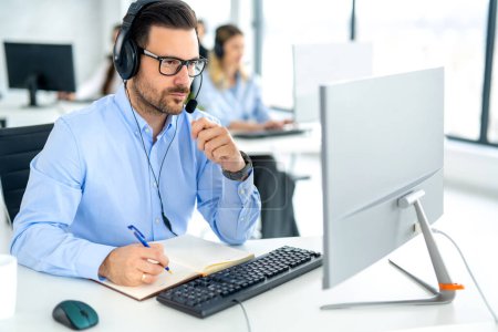 Photo for Serious successful business man wearing headset looking at computer monitor and taking notes to notebook accompanied with colleagues in the background. - Royalty Free Image