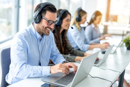 Happy male customer service assistant with headset chatting with client during online phone call and setting up customer account on laptop. Group of call center agents working on laptops.