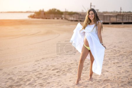 Téléchargez les photos : Portrait pleine longueur de belle femme enceinte en robe blanche posant sur la plage au coucher du soleil - en image libre de droit