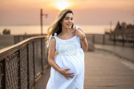 Photo for Portrait of a beautiful young pregnant woman in a white dress looking at camera on the beach promenade at sunset - Royalty Free Image