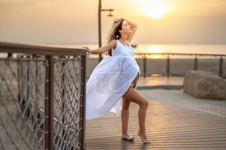 Foto de Retrato de larga duración de una hermosa mujer embarazada en vestido blanco apoyado sobre una cerca en la playa camino de madera al atardecer. - Imagen libre de derechos