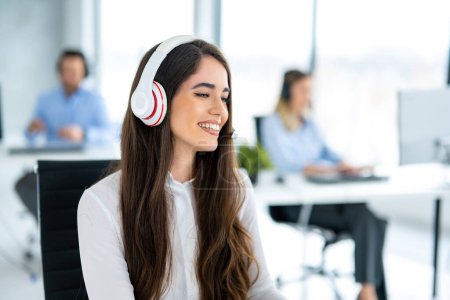 Foto de Sonriente mujer operador de línea directa con auriculares utilizando el ordenador en la oficina - Imagen libre de derechos