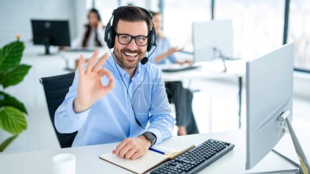 Photo for Male customer service representative showing approval sign to the camera, smiling and gesturing okay at call center office. - Royalty Free Image