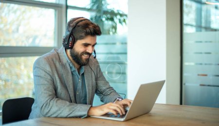 Photo for Young middle eastern businessman working on laptop computer wearing headphone at modern office. Smiling business man wearing headset for video conference, communication, education and e-learning. - Royalty Free Image