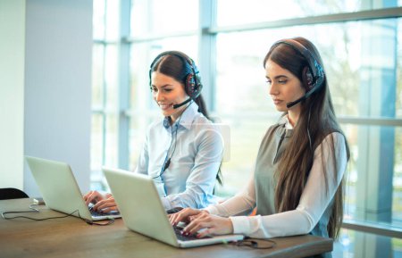 Photo for Two beautiful businesswomen at call center using laptops in corporate office. Attractive female employee workers wearing headset sitting on table using laptop, talking to customer at modern office. - Royalty Free Image