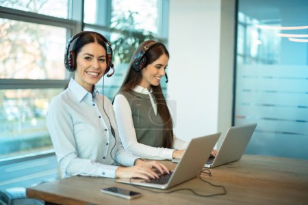 Two beautiful businesswomen at call center using laptops in corporate office. Attractive female employee workers wearing headset sitting on table using laptop, talking to support customer at workplace