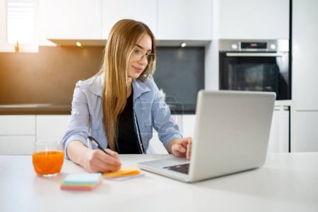 Photo for Beautiful high school student using laptop and taking notes while preparing exam or doing homework at home - Royalty Free Image
