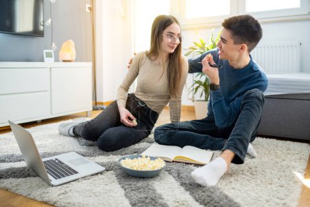 Téléchargez les photos : Jeune couple adolescent apprendre ensemble et manger du pop-corn tout en s'asseyant sur le tapis de sol embrassé à la maison - en image libre de droit