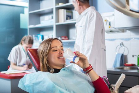 Photo for Smiling woman is taking a glass of water in dental office. Dentists in the background. - Royalty Free Image