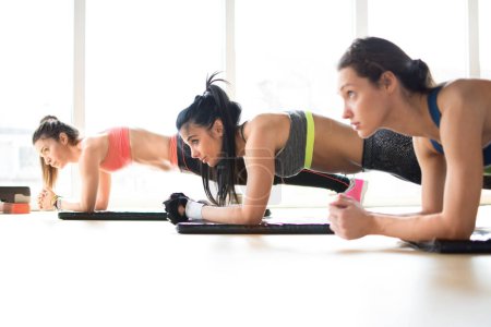 Téléchargez les photos : Trois jolies filles sportives faisant de l'exercice de planche couché sur un tapis de yoga en cours de fitness. - en image libre de droit