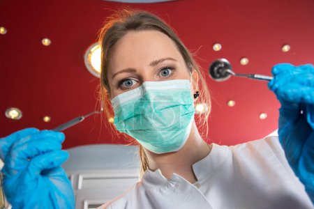 Téléchargez les photos : Vue à angle bas du dentiste féminin dans un masque chirurgical tenant des outils dentaires
. - en image libre de droit