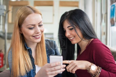 Foto de Dos chicas jóvenes usando un teléfono inteligente en la cafetería interior. - Imagen libre de derechos
