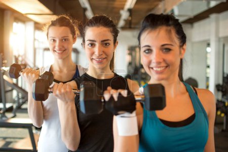Photo for Three fit and beautiful young women lifting weights in a fitness club. Focus on girl in the center. - Royalty Free Image