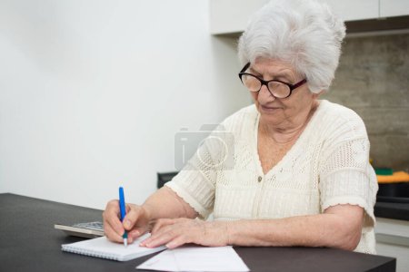 Téléchargez les photos : Femme âgée avec papiers ou factures et calculatrice écrivant à la maison. - en image libre de droit