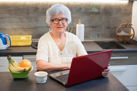 Téléchargez les photos : Femme âgée utilisant un ordinateur portable tout en faisant une pause café dans la cuisine. - en image libre de droit