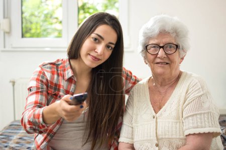 Téléchargez les photos : Petite-fille et grand-mère assises sur le canapé et regardant la télévision ensemble. - en image libre de droit