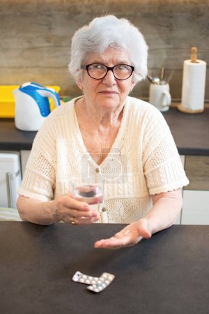 Téléchargez les photos : Portrait d'une femme âgée souriante prenant des pilules et tenant un verre d'eau à la maison. - en image libre de droit