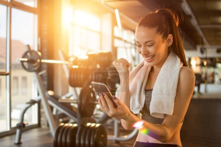 Téléchargez les photos : Femme euphorique avec beau sourire lisant de bonnes nouvelles au téléphone et célébrant le succès pendant la pause d'entraînement dans la salle de gym. - en image libre de droit