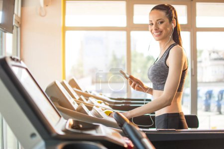 Téléchargez les photos : Femme en forme heureuse en tenue de fitness avec téléphone et écouteurs sur machine tapis roulant dans la salle de gym. - en image libre de droit