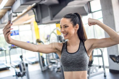 Foto de Mujer deportiva tomando una selfie con un teléfono inteligente mientras muestra su músculo del brazo en el gimnasio. - Imagen libre de derechos