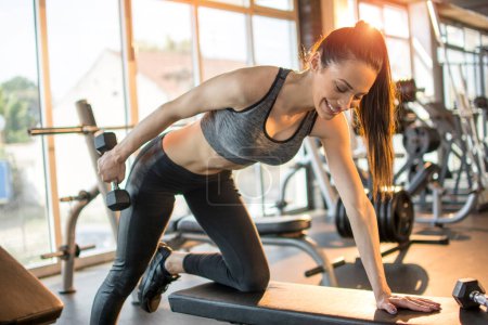 Téléchargez les photos : Souriant ajustement jeune femme flexion des muscles avec haltère dans la salle de gym. - en image libre de droit