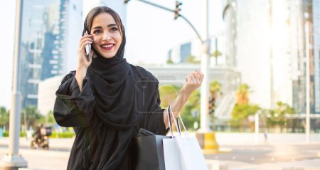 Foto de Mujer árabe en ropa tradicional sosteniendo bolsas de compras y hablando por teléfono mientras camina por la calle - Imagen libre de derechos