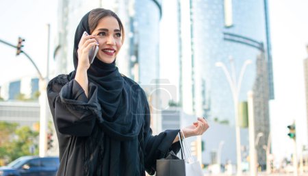 Photo for Arab woman in traditional wear holding shopping bags and talking on phone while walking on the street - Royalty Free Image