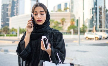 Photo for Beautiful young muslim woman in traditional clothes holding shopping bags and wallet and talking on mobile phone on the street. - Royalty Free Image