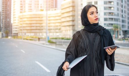 Téléchargez les photos : Belle jeune femme musulmane avec ordinateur portable et téléphone dans la rue - en image libre de droit