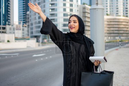 Téléchargez les photos : Jeune belle femme arabe en abaya avec des sacs à provisions, téléphone et portefeuille attelage d'un taxi dans la rue - en image libre de droit