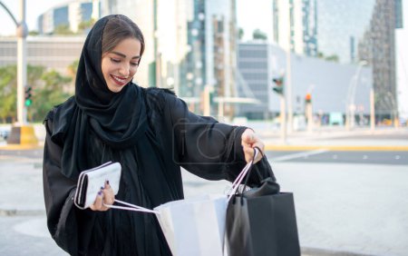 Téléchargez les photos : Belle jeune femme du Moyen-Orient regardant à l'intérieur de sacs à provisions dans la rue - en image libre de droit