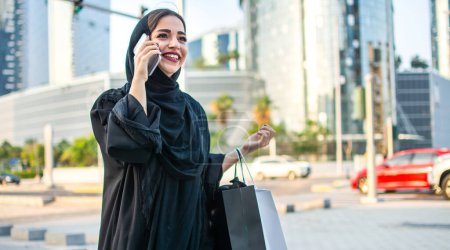 Photo for Arab woman in traditional wear holding shopping bags and talking on phone while standing on the street in front of the modern skyscrapers. - Royalty Free Image