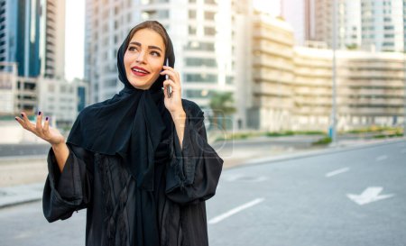 Téléchargez les photos : Jeune belle femme d'affaires arabe amicale portant Abaya parlant sur un téléphone intelligent dans la rue - en image libre de droit