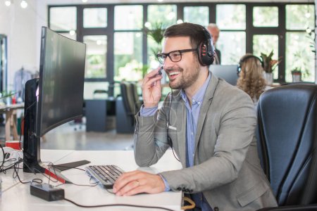 Téléchargez les photos : Opérateur de soutien à la clientèle souriant portant casque parlant avec le client tout en travaillant sur l'ordinateur dans le bureau - en image libre de droit